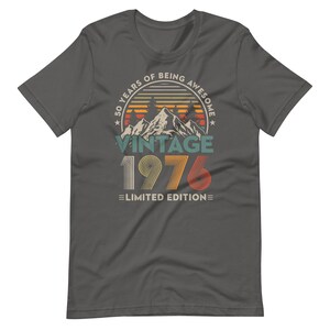 May include: Dark gray t-shirt with a vintage design. The graphic includes the text "50 YEARS OF BEING AWESOME" in a circular pattern, with "VINTAGE 1976 LIMITED EDITION" below a mountain and sunset illustration.