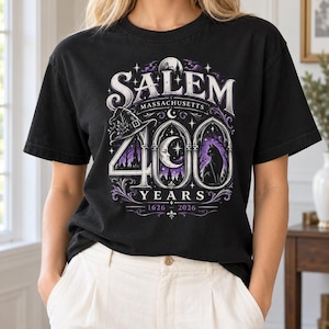 May include: A black short-sleeve t-shirt featuring a graphic print that reads SALEM MASSACHUSETTS 400 YEARS 1626-2026. The design includes a witch hat, a crescent moon, a black cat, and gothic arch motifs in shades of purple, white, and black.