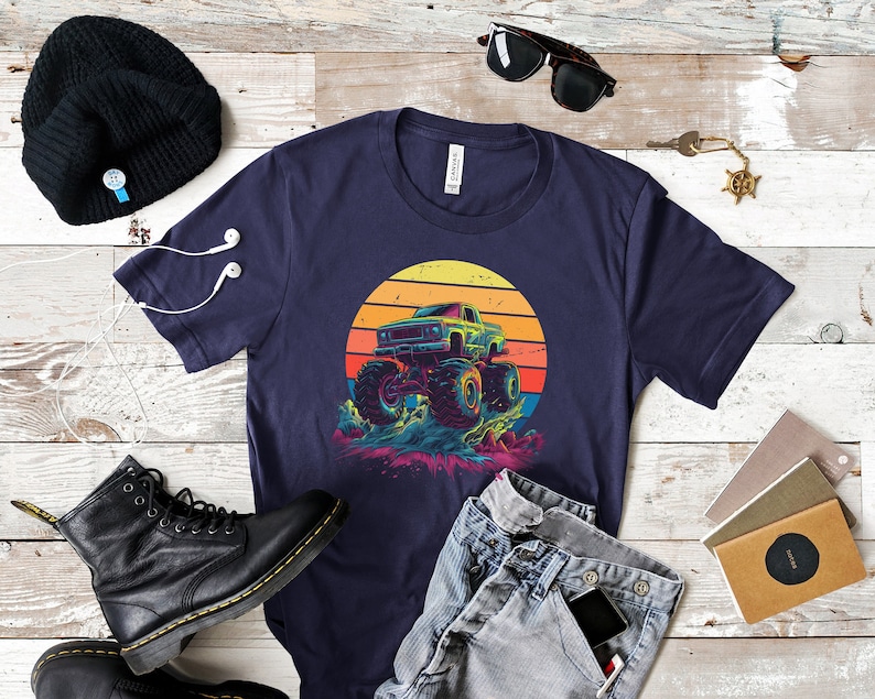 May include: A navy blue t-shirt with a graphic of a monster truck against a sunset. The design shows a detailed truck with large wheels and colourful splashes. The shirt is surrounded by accessories, including a black beanie and boots.