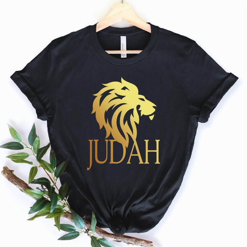 Op de afbeelding: Zwart t-shirt met een gouden leeuwenkop ontwerp en het woord "JUDAH" in gouden letters. De leeuw grafiek is gedetailleerd en gestileerd. Het shirt hangt aan een houten hanger tegen een witte achtergrond.