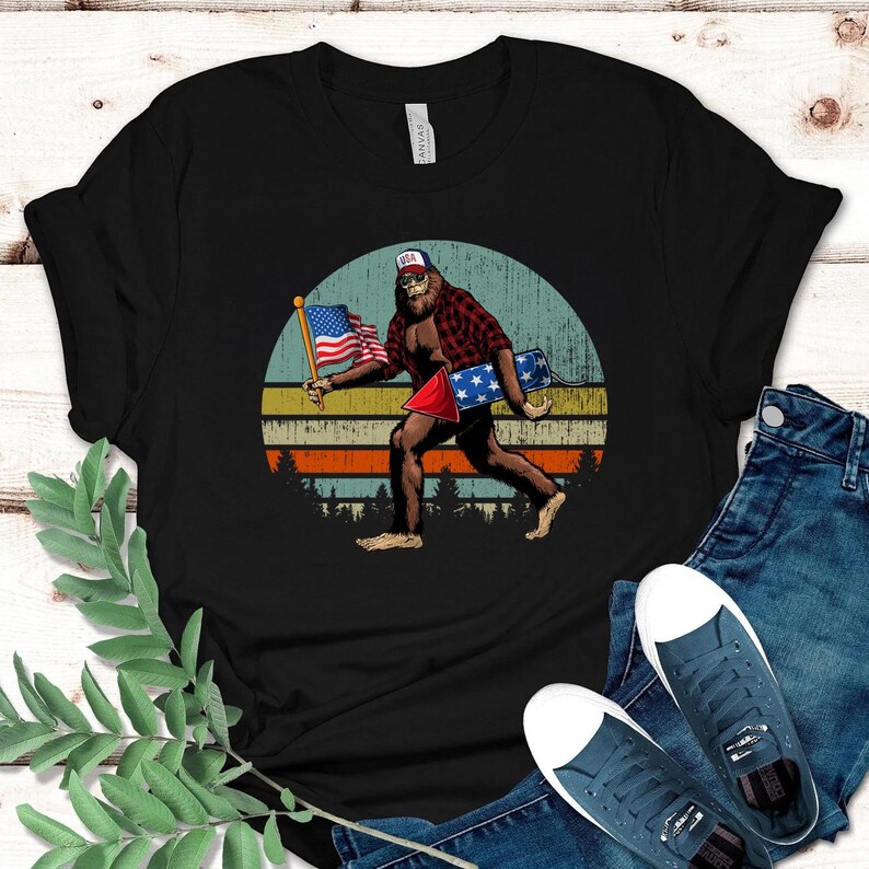 K&ouml;nnte beinhalten: Schwarzes T-Shirt mit einem Bigfoot-Motiv, das ein rot kariertes Hemd tr&auml;gt und eine amerikanische Flagge und ein Feuerwerk h&auml;lt. Das Design ist vor einem Retro-Sonnenuntergangshintergrund mit einer Waldsilhouette platziert. Der Bigfoot tr&auml;gt einen USA-Hut.