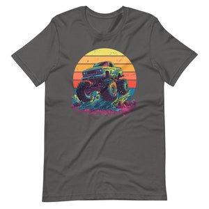 May include: Dark grey t-shirt with a colourful graphic of a monster truck jumping over a splash of water, set against a sunset. The truck is blue and green, with large tyres. The sunset has yellow, orange, and red stripes.