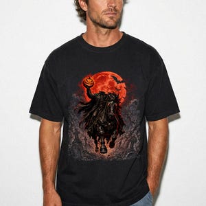 May include: A black short-sleeve t-shirt featuring a graphic print of the Headless Horseman riding a dark horse. The design includes a glowing orange full moon, flying bats, and a graveyard scene with a pumpkin held by the rider.