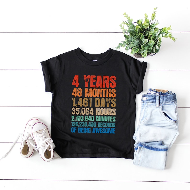 May include: Black t-shirt with the text "4 YEARS, 48 MONTHS, 1,461 DAYS, 35,064 HOURS, 2,103,840 MINUTES, 126,230,400 SECONDS OF BEING AWESOME" in a retro style. White sneakers and blue jeans are next to the shirt.