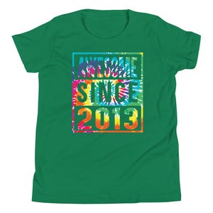 May include: A green t-shirt featuring the text "AWESOME SINCE 2013" in a tie-dye design. The text is in a rectangular shape with blue, pink, yellow, and orange colours. The shirt is a solid green colour.