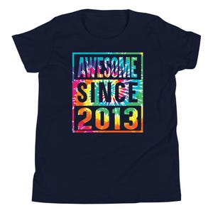 May include: Navy blue t-shirt with a colourful tie-dye design. The shirt features the text "AWESOME SINCE 2013" in a rectangular frame. The text is in white, yellow, orange, and blue. The shirt is a youth size.