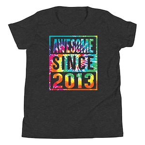 May include: Dark grey t-shirt with a vibrant tie-dye pattern. The shirt displays the text "AWESOME SINCE 2013" within a rectangular border. The text is black, and the tie-dye includes blue, green, yellow, orange, and red.