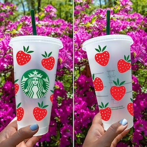 Puede incluir: Un vaso reutilizable de plástico transparente con un logotipo de Starbucks verde y diseños de fresas rojas. El vaso tiene una pajita verde y está siendo sostenido por una mano.