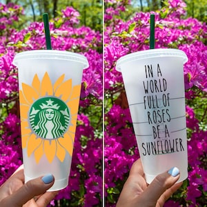 Può includere: Due bicchieri riutilizzabili in plastica trasparente con coperchi e cannucce. Un bicchiere presenta un design a girasole con un logo verde Starbucks al centro. L'altro bicchiere ha la scritta "In un mondo pieno di rose, sii un girasole" stampata su di esso.