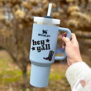 May include: A light blue stainless steel travel mug with a black handle and a black and white graphic that says "hey yall" with a cowboy boot and a star. The mug has a black Stanley logo and a black straw.