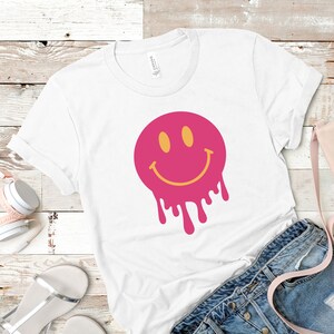 Puede incluir: Camiseta blanca con un gráfico de cara sonriente rosa que gotea. La cara sonriente tiene un contorno amarillo y ojos amarillos.