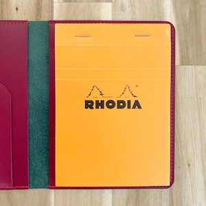 May include: A burgundy leather notebook cover with a green spine and an orange notepad. The notepad has the brand name "RHODIA" printed in black. The cover is open, revealing the notepad and interior pockets.