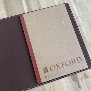 Può includere: Quaderno in pelle marrone aperto con un inserto di carta a righe. La carta ha un dorso rosso e la parola "OXFORD" stampata in marrone. Il quaderno è aperto su una superficie di legno chiaro.