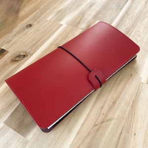 Op de afbeelding: Een rood leren notitieboek met een zwarte elastische sluiting. Het notitieboek is rechthoekig en heeft een gestikte rand. De omslag is effen, levendig rood. Het notitieboek is gesloten met een zwarte elastische band.