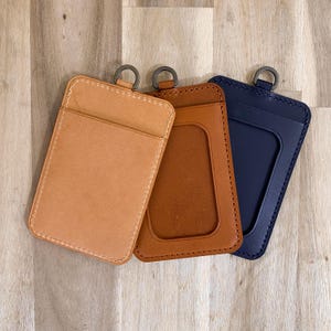 May include: Three leather ID badge holders in tan, brown, and navy blue. Each holder has a metal ring at the top and a stitched border. The brown holder has a clear window for displaying an ID.