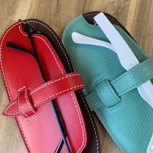 Może przedstawiać: Dwa skórzane etui na okulary. Jedno jest czerwone z białym obszyciem i pasującym paskiem. Drugie jest turkusowe z białym obszyciem i pasującym paskiem. Czarne okulary znajdują się w czerwonym etui, a białe przybory w turkusowym etui.