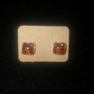May include: A pair of amber earrings with a square shape and a silver setting. The earrings are displayed on a brown card.