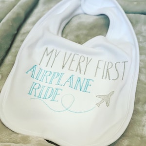 May include: White baby bib with the text "My Very First Airplane Ride" embroidered in gray and blue. A small airplane with a blue tail is embroidered next to the text.
