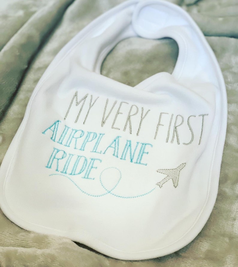 May include: White baby bib with the text "My Very First Airplane Ride" embroidered in gray and blue. A small airplane with a blue tail is embroidered next to the text.