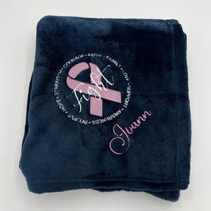 May include: A navy blue fleece blanket with a pink ribbon and the word "fight" embroidered in white. The words "believe", "hope", "strength", "courage", "faith", "family", "love", "support", "awareness" and the name "Joann" are also embroidered in white around the ribbon.