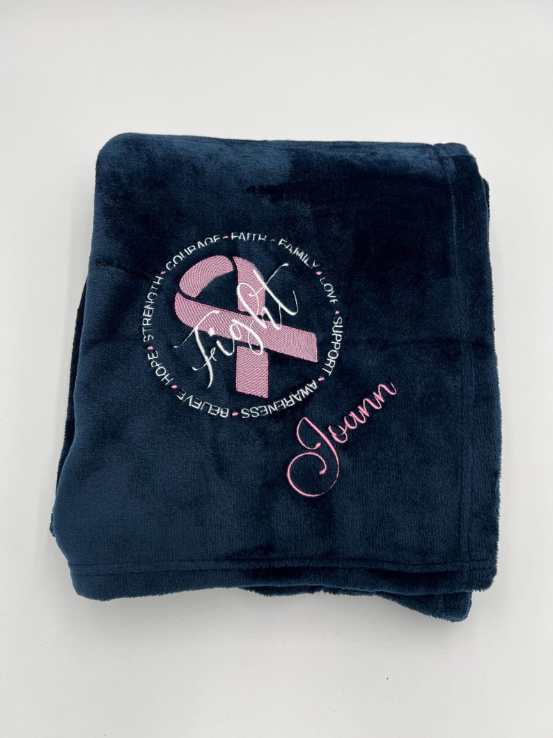 May include: A navy blue fleece blanket with a pink ribbon and the word "fight" embroidered in white. The words "believe", "hope", "strength", "courage", "faith", "family", "love", "support", "awareness" and the name "Joann" are also embroidered in white around the ribbon.