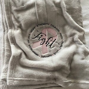 May include: A gray fleece blanket with a pink ribbon and the word "fight" embroidered in a circle. The circle also includes the words "believe", "hope", "strength", "courage", "faith", "family", "love", "support", and "awareness".