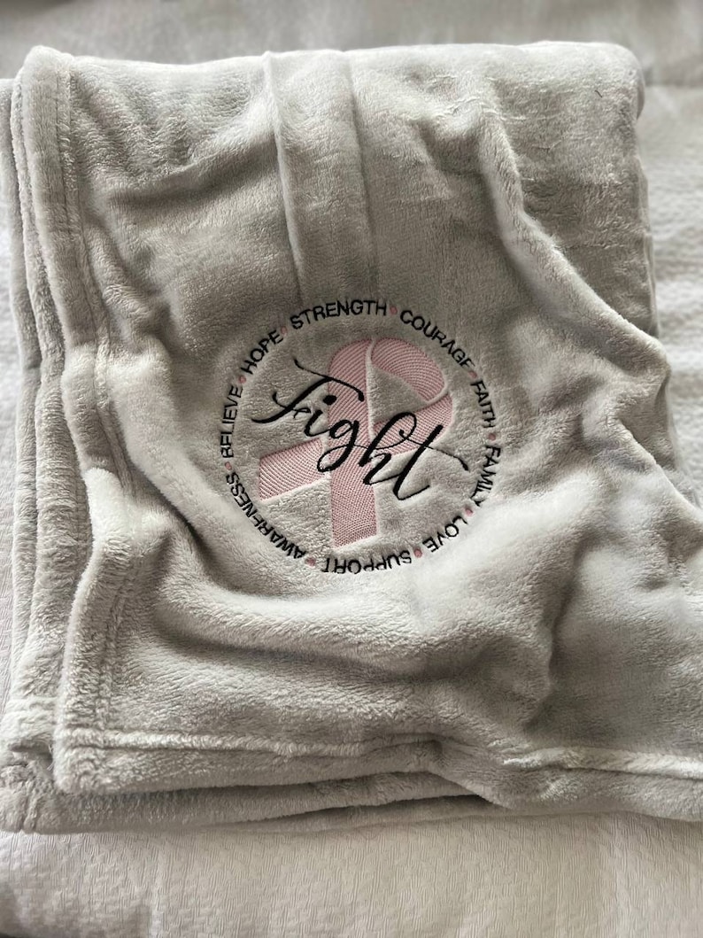 May include: A gray fleece blanket with a pink ribbon and the word "fight" embroidered in a circle. The circle also includes the words "believe", "hope", "strength", "courage", "faith", "family", "love", "support", and "awareness".