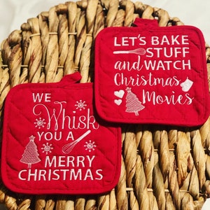 May include: Two red quilted pot holders with white embroidered Christmas-themed sayings. The first pot holder says "We Whisk You A Merry Christmas" with a Christmas tree, snowflakes, and a whisk. The second pot holder says "Let's Bake Stuff and Watch Christmas Movies" with a Christmas tree, hearts, and a whisk.