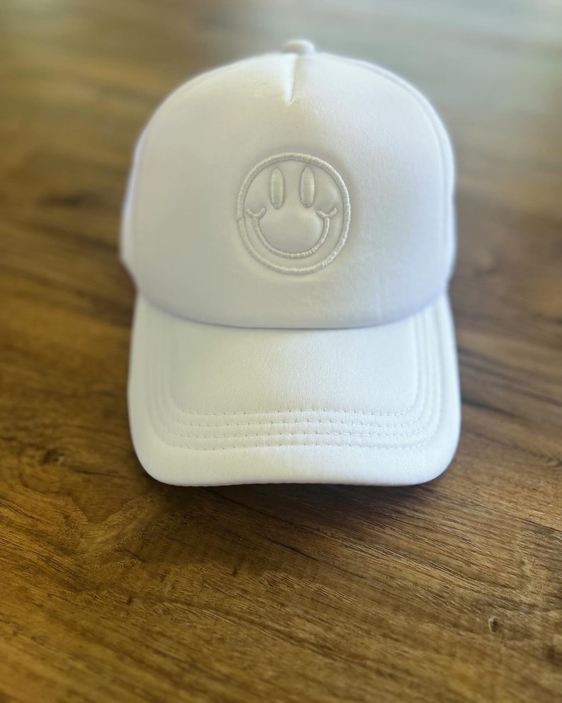 K&ouml;nnte beinhalten: Wei&szlig;e Trucker-Cap mit einem Smiley-Aufn&auml;her. Die Kappe hat einen gebogenen Schirm und eine Mesh-R&uuml;ckseite. Das Smiley-Gesicht ist auf der Vorderseite aufgestickt. Die Kappe besteht aus einem weichen, atmungsaktiven Material.
