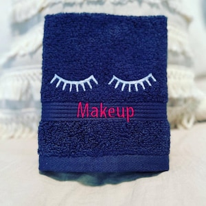 May include: A navy blue face towel with white embroidered eyelashes and the word "Makeup" in pink. The towel is rectangular and made of a soft, absorbent material, perfect for removing makeup.