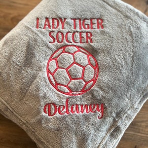 May include: A gray, plush blanket embroidered with "LADY TIGER SOCCER", a red soccer ball, and the name "Delaney" in red thread. The blanket is folded and appears soft and cozy.