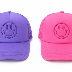 K&ouml;nnte beinhalten: Zwei Baseballkappen, eine lila und eine rosa, jeweils mit einem Smiley-Gesicht-Design. Die Kappen haben einen Mesh-R&uuml;cken und einen gebogenen Schirm. Das Smiley-Gesicht ist auf der Vorderseite jeder Kappe gestickt.