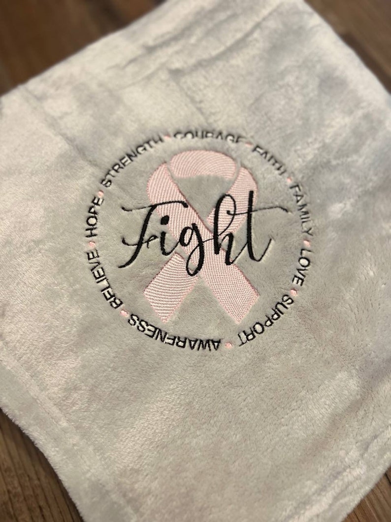 May include: White plush blanket with a pink ribbon and the word "Fight" embroidered in the center. The ribbon is surrounded by a circle of words including "Strength", "Courage", "Faith", "Family", "Love", "Support", "Awareness", "Believe", and "Hope".