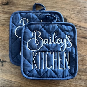 May include: Two blue quilted pot holders with white embroidery. The text on the pot holders reads "Bailey's Kitchen".
