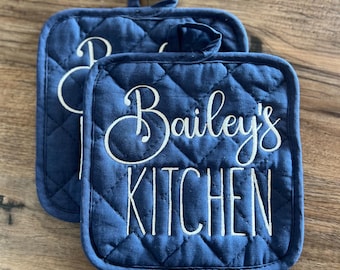 May include: Two blue quilted pot holders with white embroidery. The text on the pot holders reads "Bailey's Kitchen".