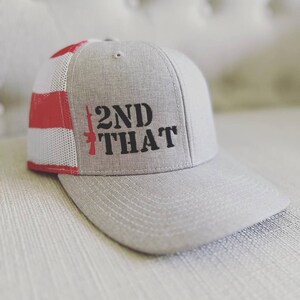 May include: A gray and red trucker hat with a white mesh back. The hat has a black embroidered design that says "2ND THAT" with a stylized rifle graphic.