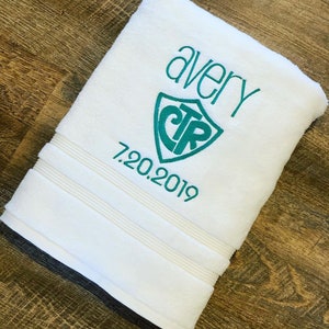 May include: White bath towel with teal embroidery. The embroidery includes the name "avery", a shield with the letters "CTR" inside, and the date "7.20.2019".