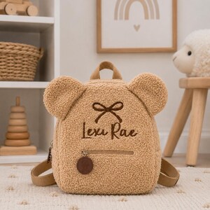 Puede incluir: Una mochila en forma de oso de peluche de color beige con el nombre "Lexi Rae" bordado en la parte delantera. La mochila tiene un peque&ntilde;o bolsillo con cremallera, correas ajustables y un asa superior. Las orejas son una caracter&iacute;stica destacada.