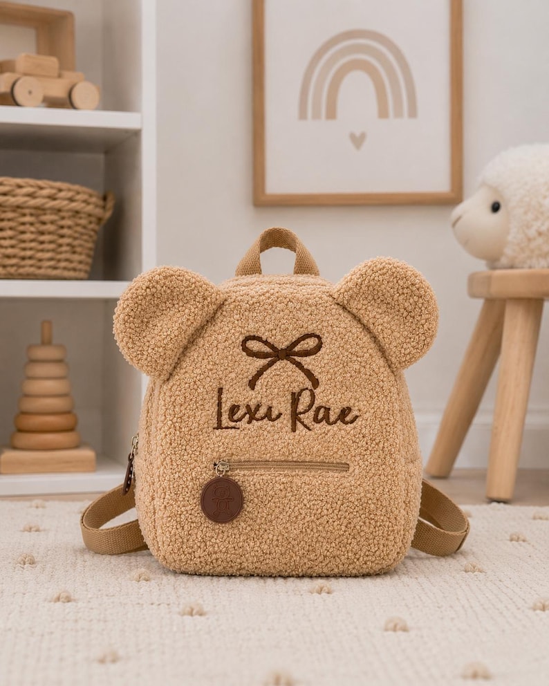 Puede incluir: Una mochila en forma de oso de peluche de color beige con el nombre "Lexi Rae" bordado en la parte delantera. La mochila tiene un peque&ntilde;o bolsillo con cremallera, correas ajustables y un asa superior. Las orejas son una caracter&iacute;stica destacada.