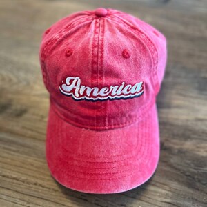 Puede incluir: Una gorra de b&eacute;isbol de algod&oacute;n roja con un "America" bordado en blanco en una fuente retro.