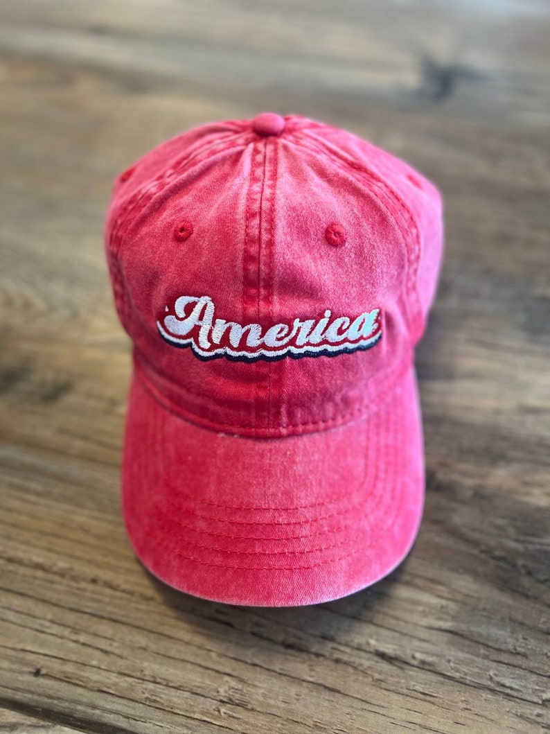 Puede incluir: Una gorra de b&eacute;isbol de algod&oacute;n roja con un "America" bordado en blanco en una fuente retro.