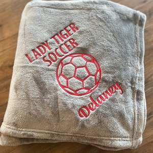May include: A gray, plush blanket embroidered with "LADY TIGER SOCCER", a soccer ball, and the name "Delaney" in red thread. The blanket is folded and appears soft and cozy.