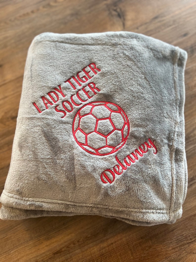 May include: A gray, plush blanket embroidered with "LADY TIGER SOCCER", a soccer ball, and the name "Delaney" in red thread. The blanket is folded and appears soft and cozy.