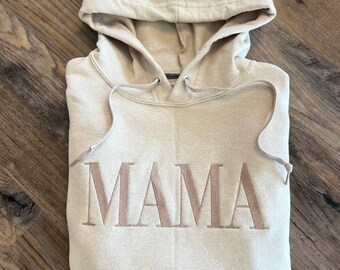 May include: A beige hooded sweatshirt with the word "MAMA" embroidered in brown on the front.