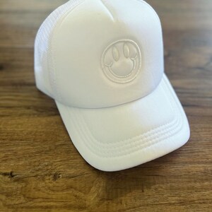K&ouml;nnte beinhalten: Wei&szlig;e Trucker-Cap mit einem Smiley-Aufn&auml;her. Die Kappe hat eine Mesh-R&uuml;ckseite und einen gebogenen Schirm. Das Smiley-Gesicht ist auf der Vorderseite aufgestickt. Der Hut ist aus einem weichen, atmungsaktiven Material gefertigt.