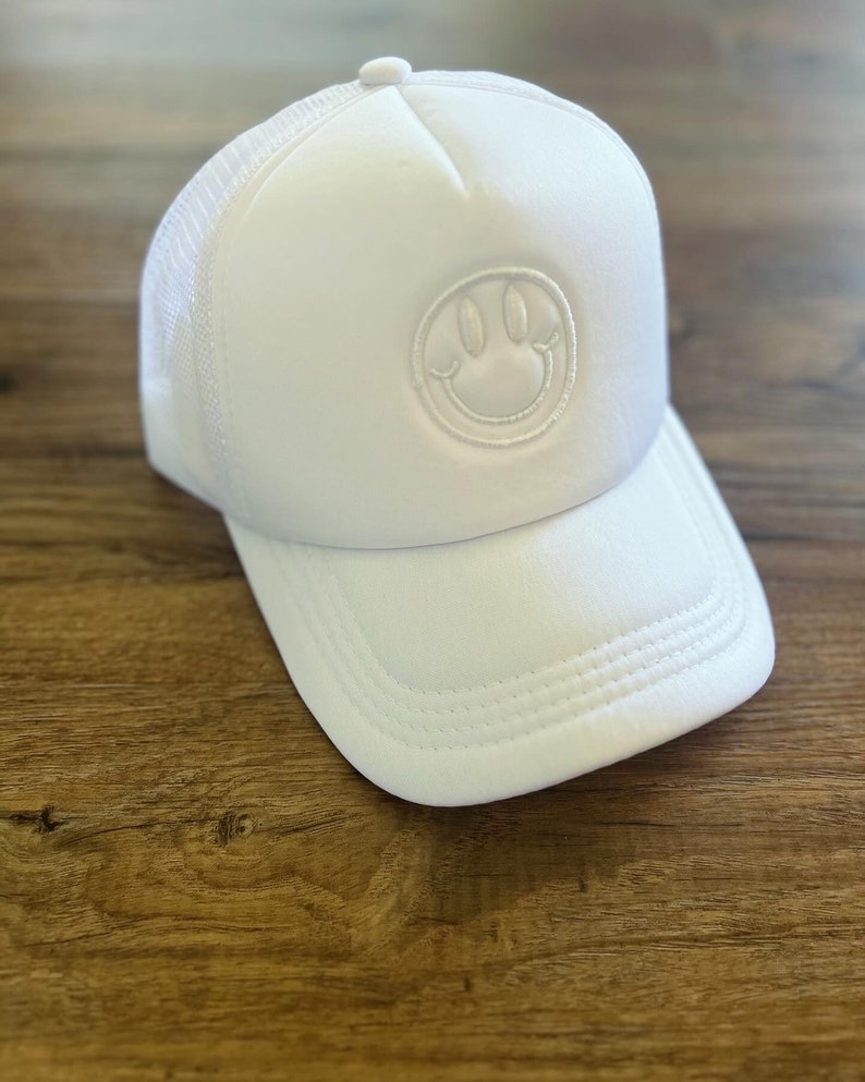 K&ouml;nnte beinhalten: Wei&szlig;e Trucker-Cap mit einem Smiley-Aufn&auml;her. Die Kappe hat eine Mesh-R&uuml;ckseite und einen gebogenen Schirm. Das Smiley-Gesicht ist auf der Vorderseite aufgestickt. Der Hut ist aus einem weichen, atmungsaktiven Material gefertigt.