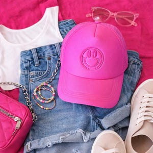 K&ouml;nnte beinhalten: Draufsicht auf ein l&auml;ssiges Outfit auf rosa Hintergrund. Das Outfit beinhaltet ein wei&szlig;es Tanktop, Jeansshorts, eine pinkfarbene Baseballkappe mit Smiley, eine pinkfarbene Sonnenbrille, eine pinkfarbene Tasche, eine Perlenkette und beige Sneaker.