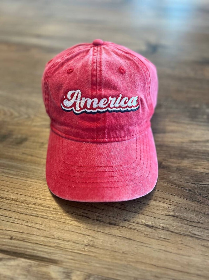 Puede incluir: Una gorra de b&eacute;isbol roja con la palabra "America" bordada en blanco en la parte delantera. La gorra tiene un aspecto desgastado y una visera curvada.