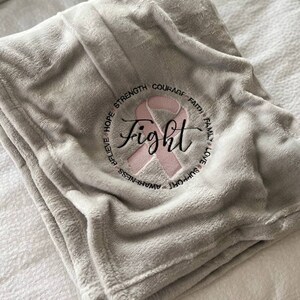 May include: A gray fleece blanket with a pink ribbon and the word "fight" embroidered in black. The ribbon is surrounded by the words "support, awareness, believe, hope, strength, courage, faith, family, love".