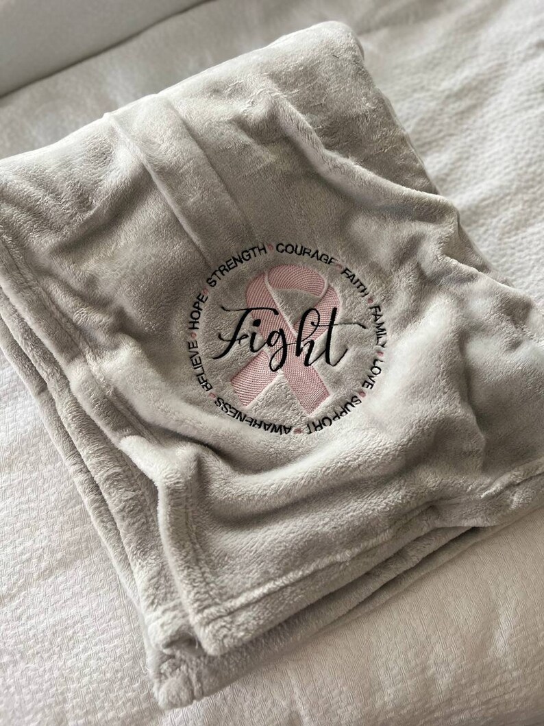 May include: A gray fleece blanket with a pink ribbon and the word "fight" embroidered in black. The ribbon is surrounded by the words "support, awareness, believe, hope, strength, courage, faith, family, love".
